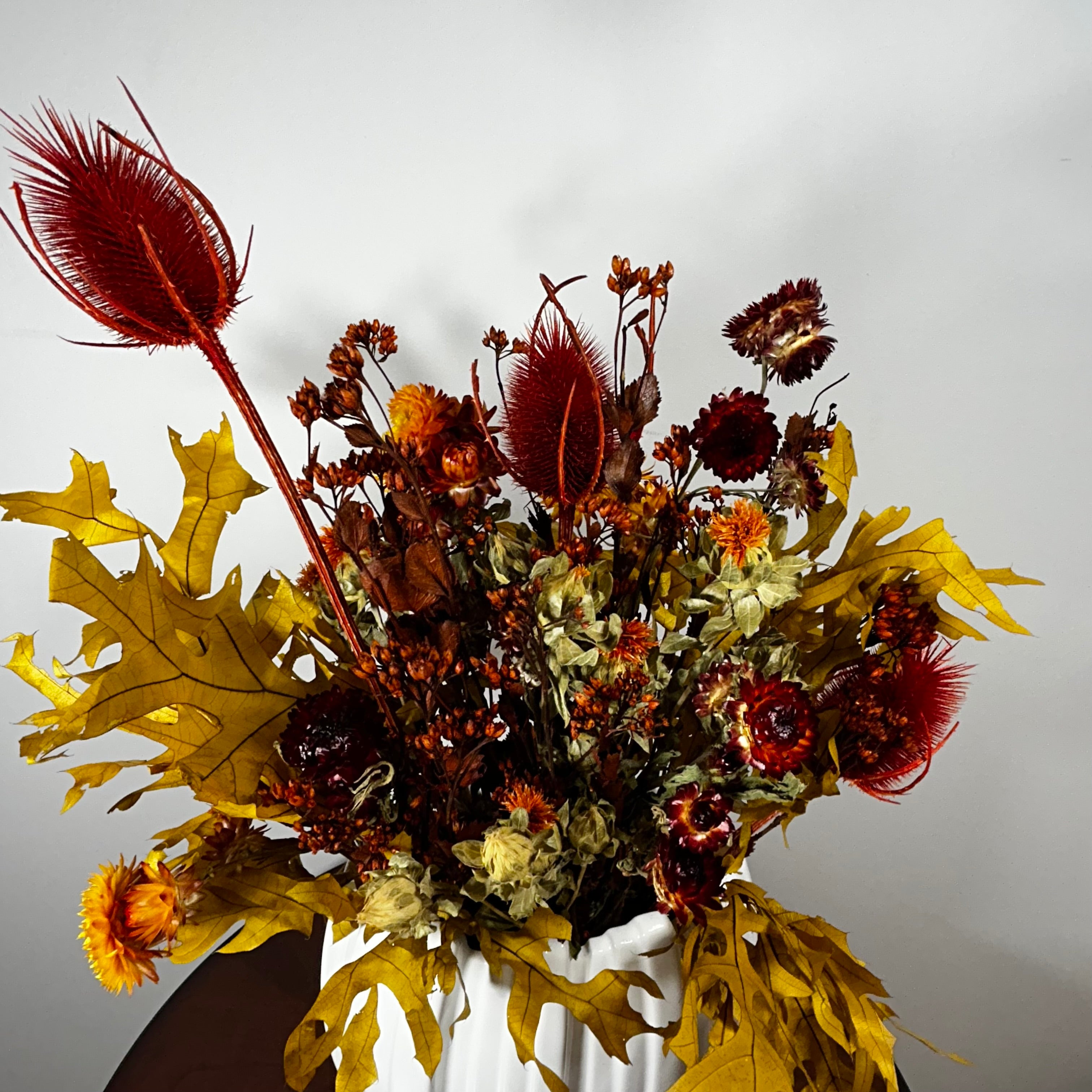 A dried floral arrangement in a white ceramic vase, featuring golden oak leaves, red feathery stems, and mixed autumn flowers against a plain light gray wall.