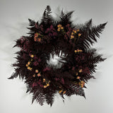 A circular floral wreath featuring dark burgundy ferns, clusters of small pink flowers, and scattered pale peach dried blossoms, photographed against a plain light gray background.