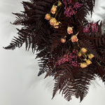A dried floral arrangement featuring dark burgundy ferns, small pink flowers, and pale yellow roses, photographed against a plain light gray background.