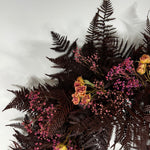 A dried floral arrangement featuring dark burgundy ferns, pink baby's breath, and dried roses, photographed from an overhead angle against a light gray background.