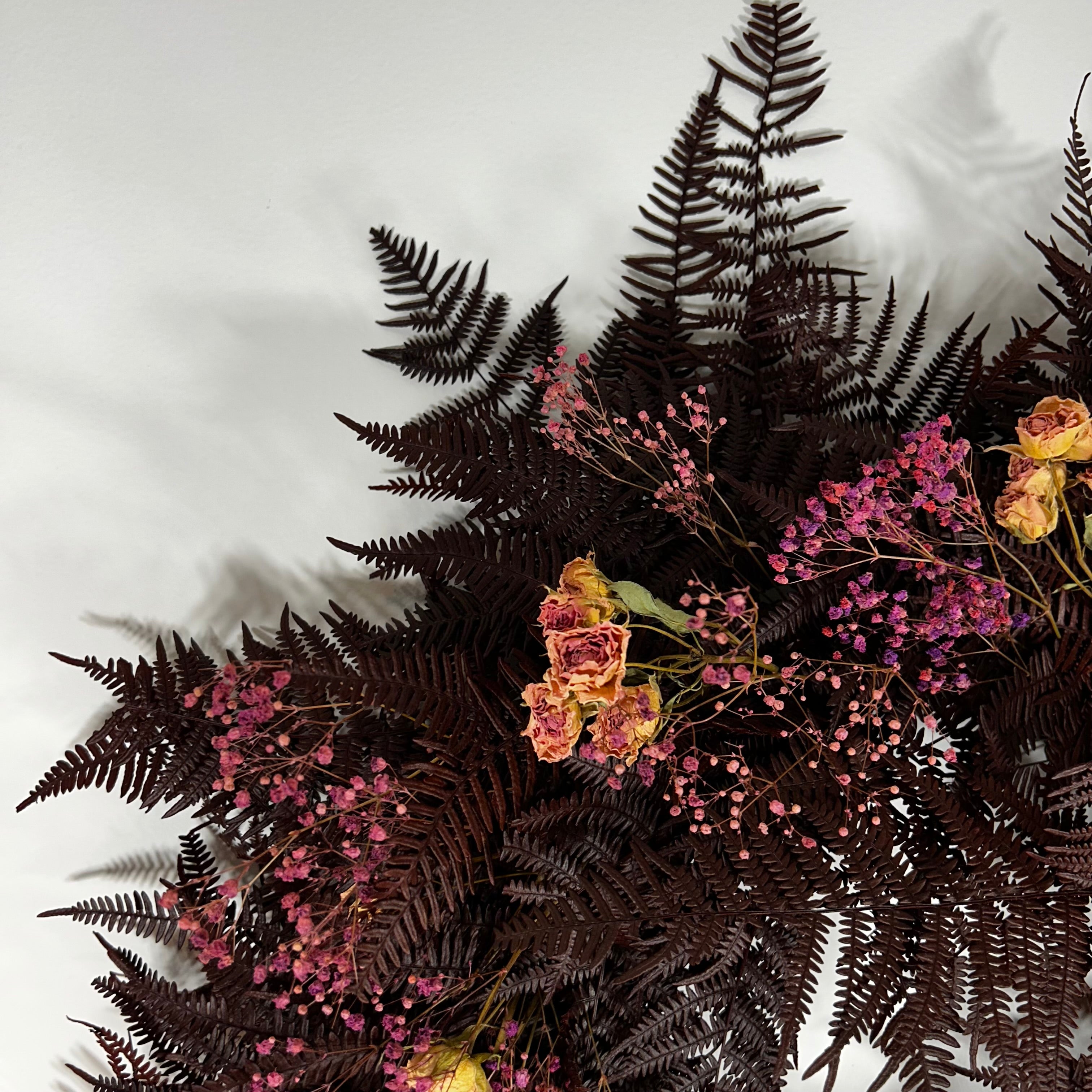 A dried floral arrangement featuring dark burgundy ferns, pink baby's breath, and dried roses, photographed from an overhead angle against a light gray background.