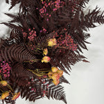 A dried floral arrangement featuring dark burgundy ferns, small pink flowers, and dried yellow roses on a plain white background.