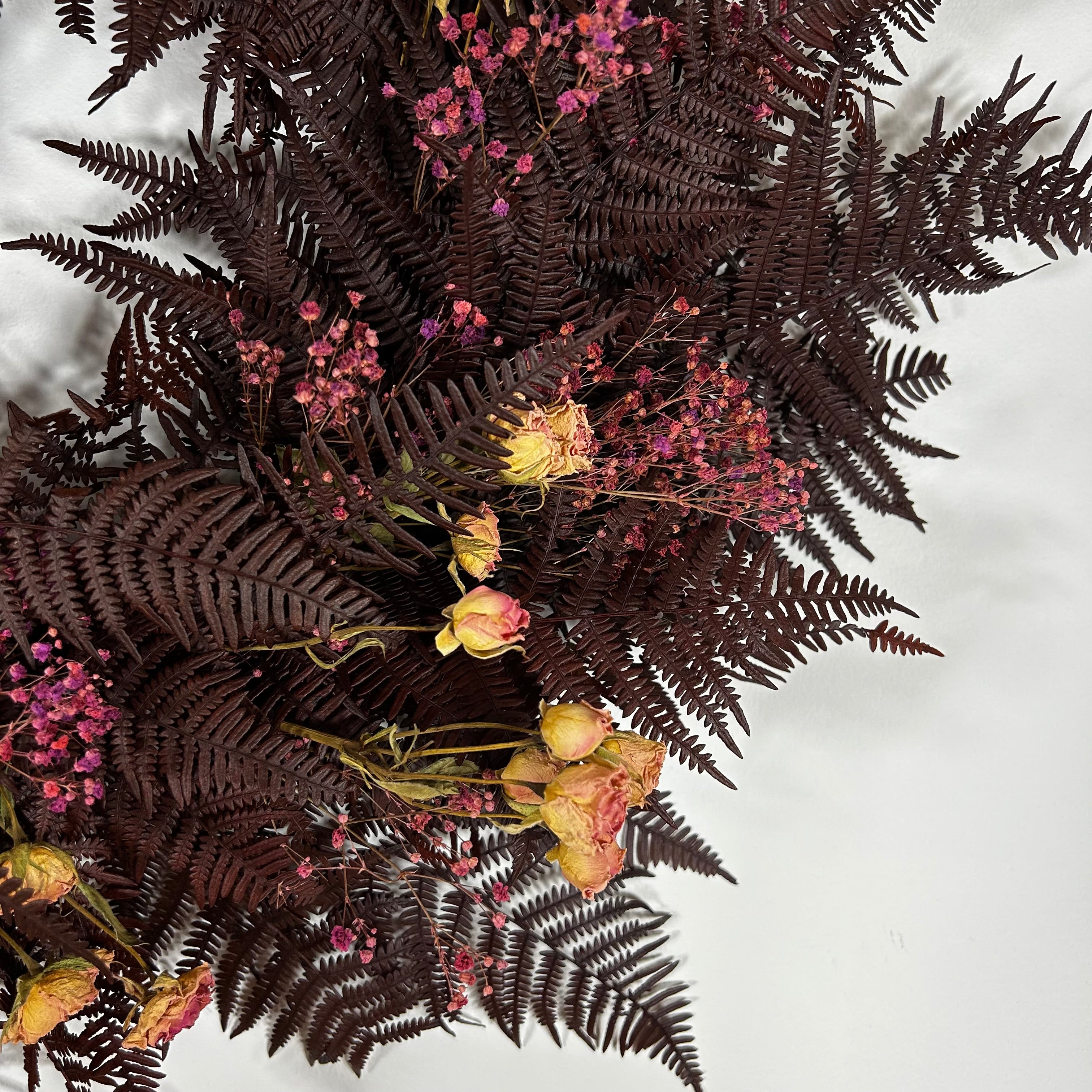 A dried floral arrangement featuring dark burgundy ferns, small pink flowers, and dried yellow roses on a plain white background.