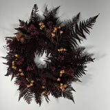 A dark, moody dried floral wreath featuring prominent dark brown ferns, clusters of small dusty pink flowers, and scattered pale yellow blooms, set against a plain light gray background.