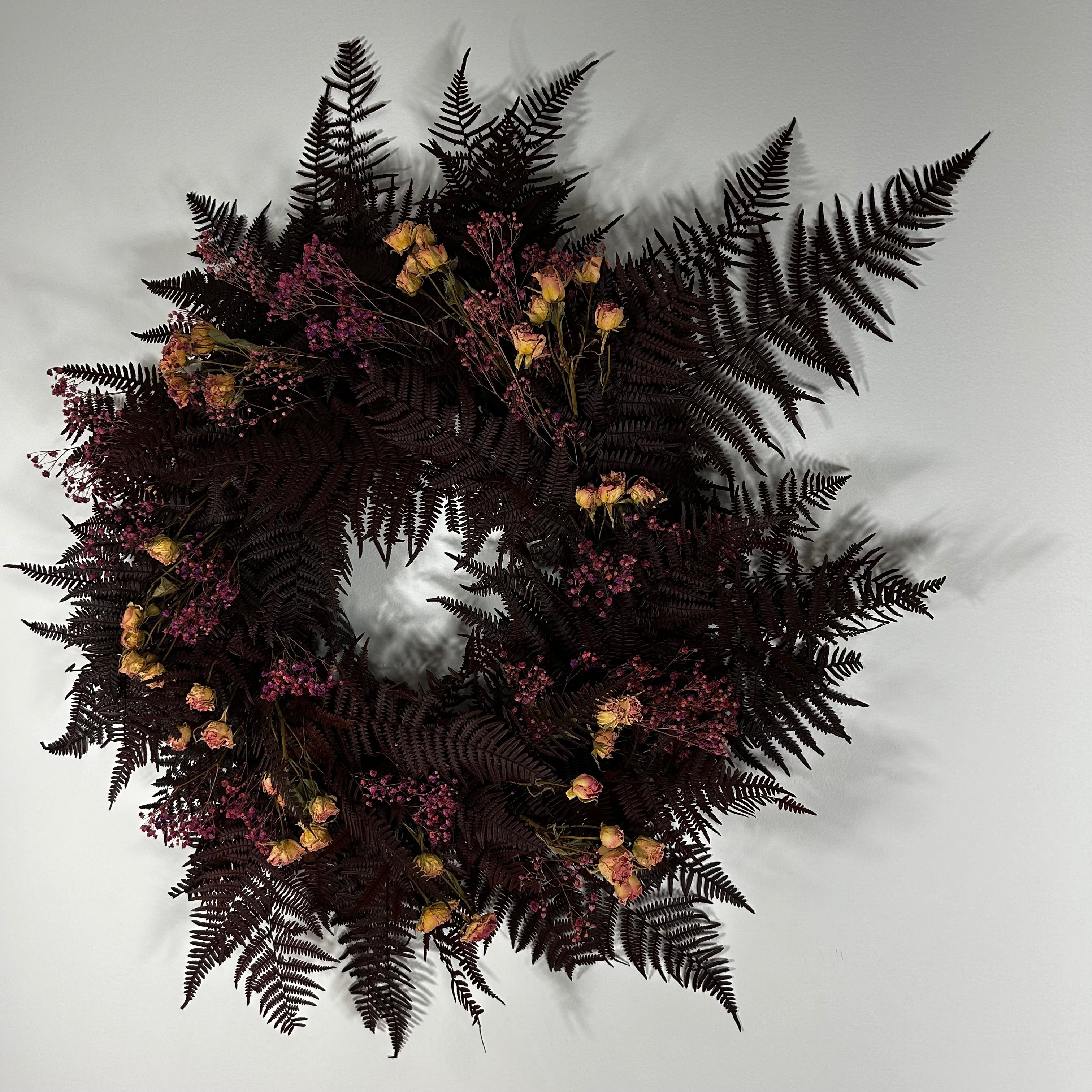 A dark, moody dried floral wreath featuring prominent dark brown ferns, clusters of small dusty pink flowers, and scattered pale yellow blooms, set against a plain light gray background.