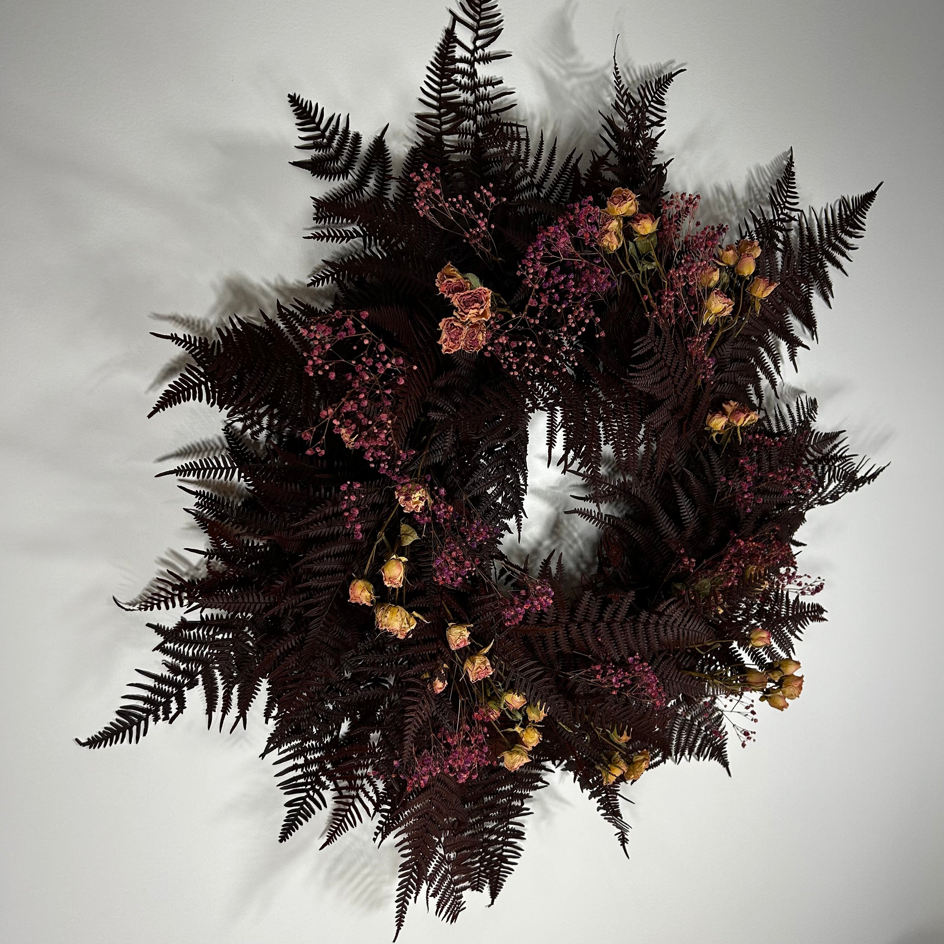 A dark, moody floral wreath featuring deep burgundy ferns, clusters of tiny pinkish-purple flowers, and dried peach-orange blossoms, shown against a plain light gray background.