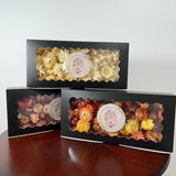Three black gift boxes containing dried flowers, displayed on a dark wooden table against a plain white wall. Each box has a scalloped clear window showing the colorful dried flowers inside, with a round white label in the center.