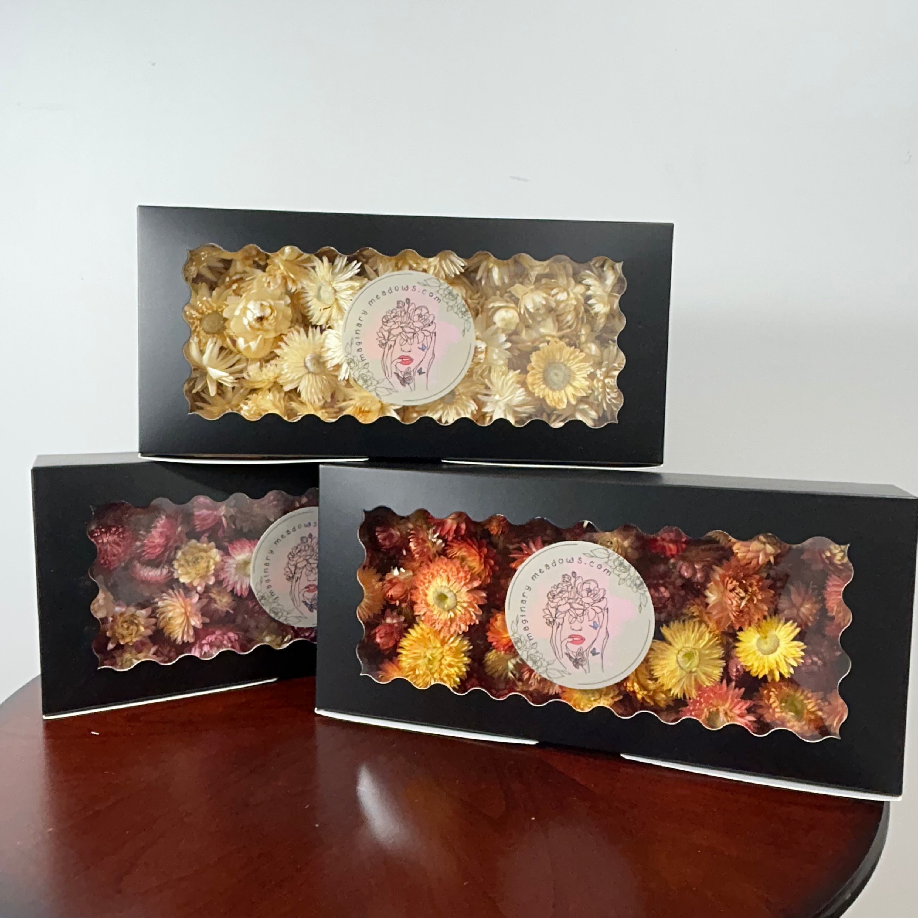 Three black gift boxes containing dried flowers, displayed on a dark wooden table against a plain white wall. Each box has a scalloped clear window showing the colorful dried flowers inside, with a round white label in the center.