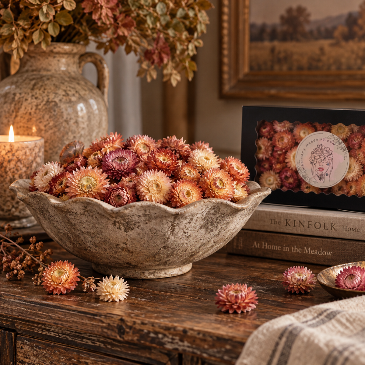 A rustic stone bowl filled with dried strawflowers in warm pink, peach, and cream tones, styled on a wooden table beside a candle, books, a framed print, and a decorative vase.