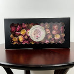 A rectangular black gift box with a scalloped clear window displaying dried flowers in pink, burgundy, and yellow tones. A round white sticker with a floral face illustration and the text 'Dried Flower Fragrance' is attached to the front.
