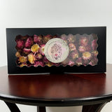 A rectangular black gift box with a scalloped clear window displaying dried flowers in pink, burgundy, and yellow tones. A round white sticker with a floral face illustration and the text 'Dried Flower Fragrance' is attached to the front.