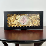 A black rectangular gift box with a clear scalloped window displaying a cream coloured dried flowers and a round decorative label, set on a dark wooden table against a light gray wall.
