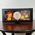 A rectangular black gift box with a scalloped clear window displaying dried multicolored flowers, with a round white label in the center. The box sits on a dark wooden table against a plain light gray wall.