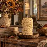 An ornate cream-colored pillar candle with raised sunflower motifs sits on a rustic wooden table in a warm, decorated room. The candle is displayed on a matching metal tray, surrounded by autumn-themed decor including a vase of flowers, books, apples, and a small bird figurine.