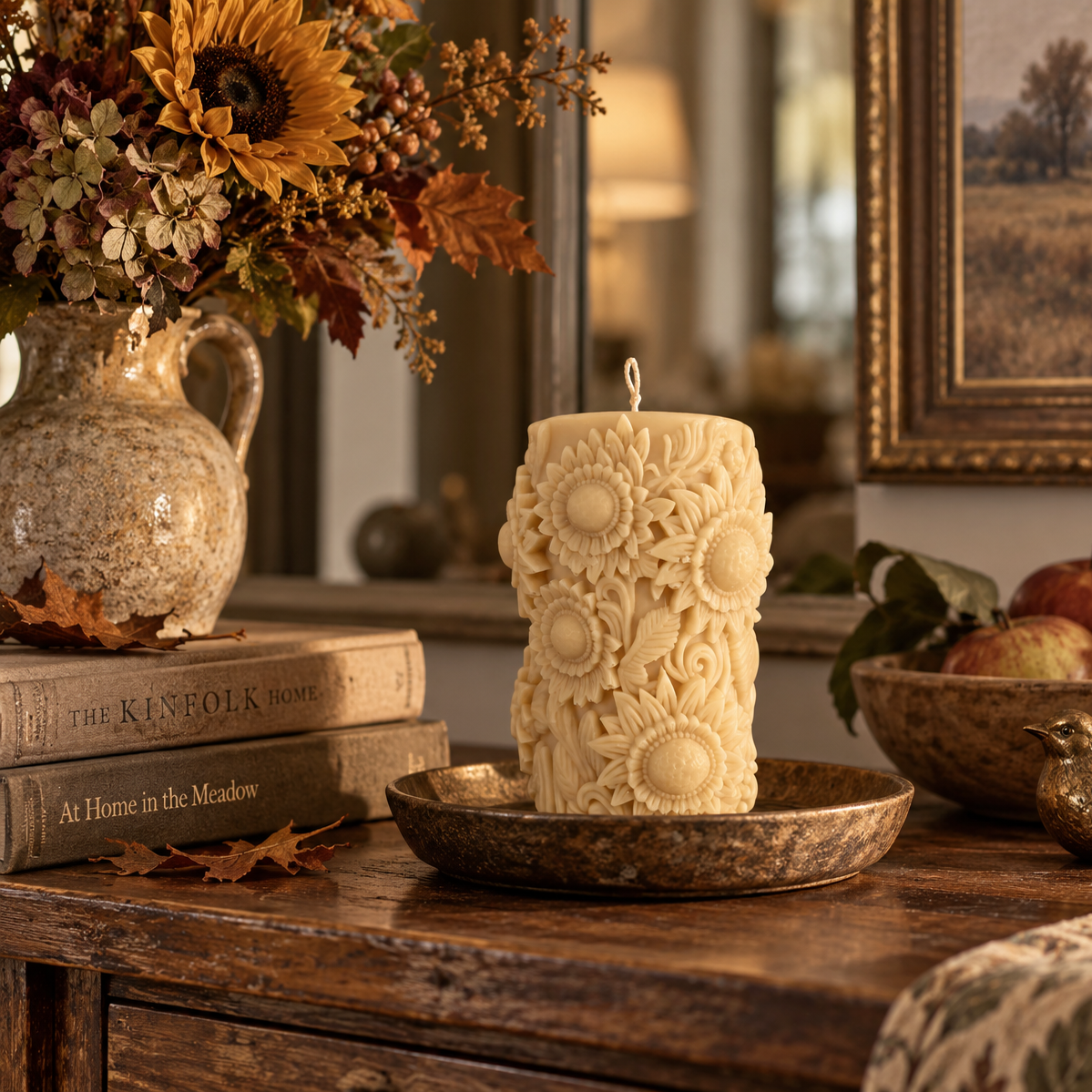 An ornate cream-colored pillar candle with raised sunflower motifs sits on a rustic wooden table in a warm, decorated room. The candle is displayed on a matching metal tray, surrounded by autumn-themed decor including a vase of flowers, books, apples, and a small bird figurine.