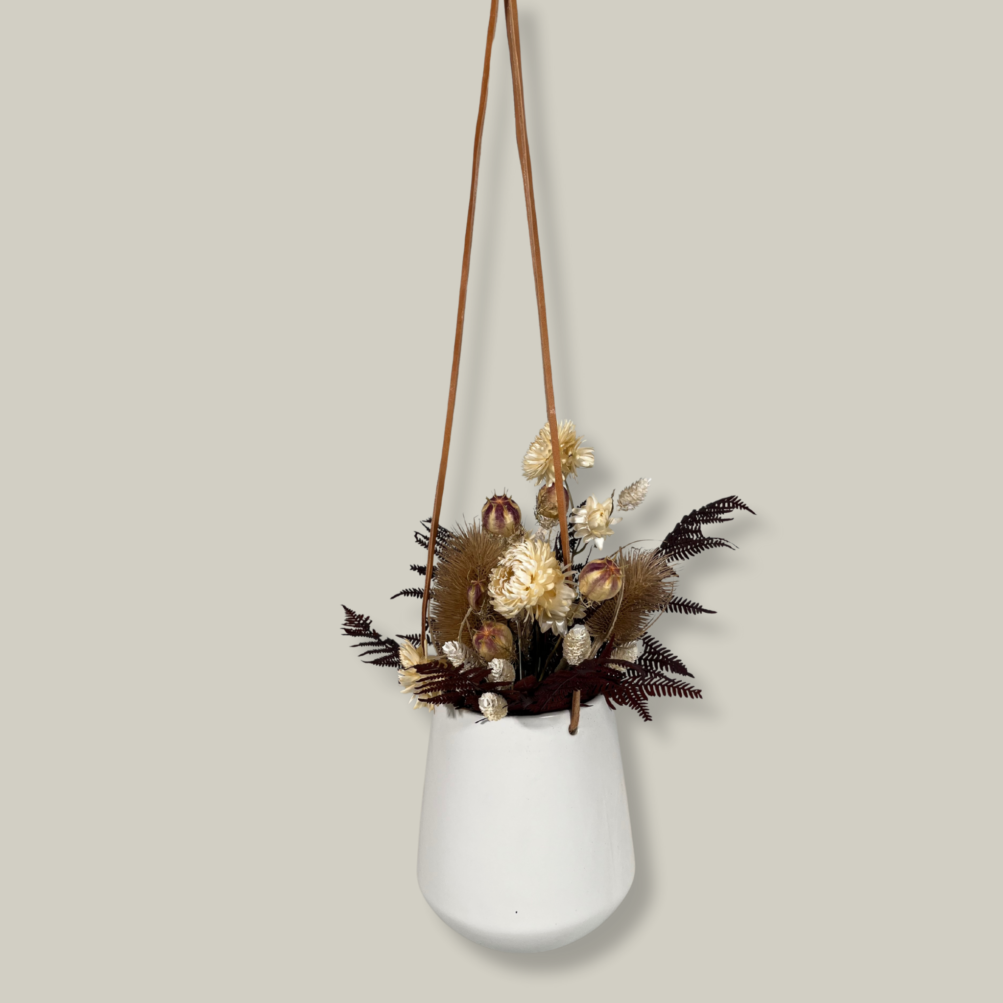 White hanging planter with beautiful dried flowers arrangement with burgundy bracken, ivory helichrysums, Nigel seed pods, natural teasels, and ivory phalaris on a beige background