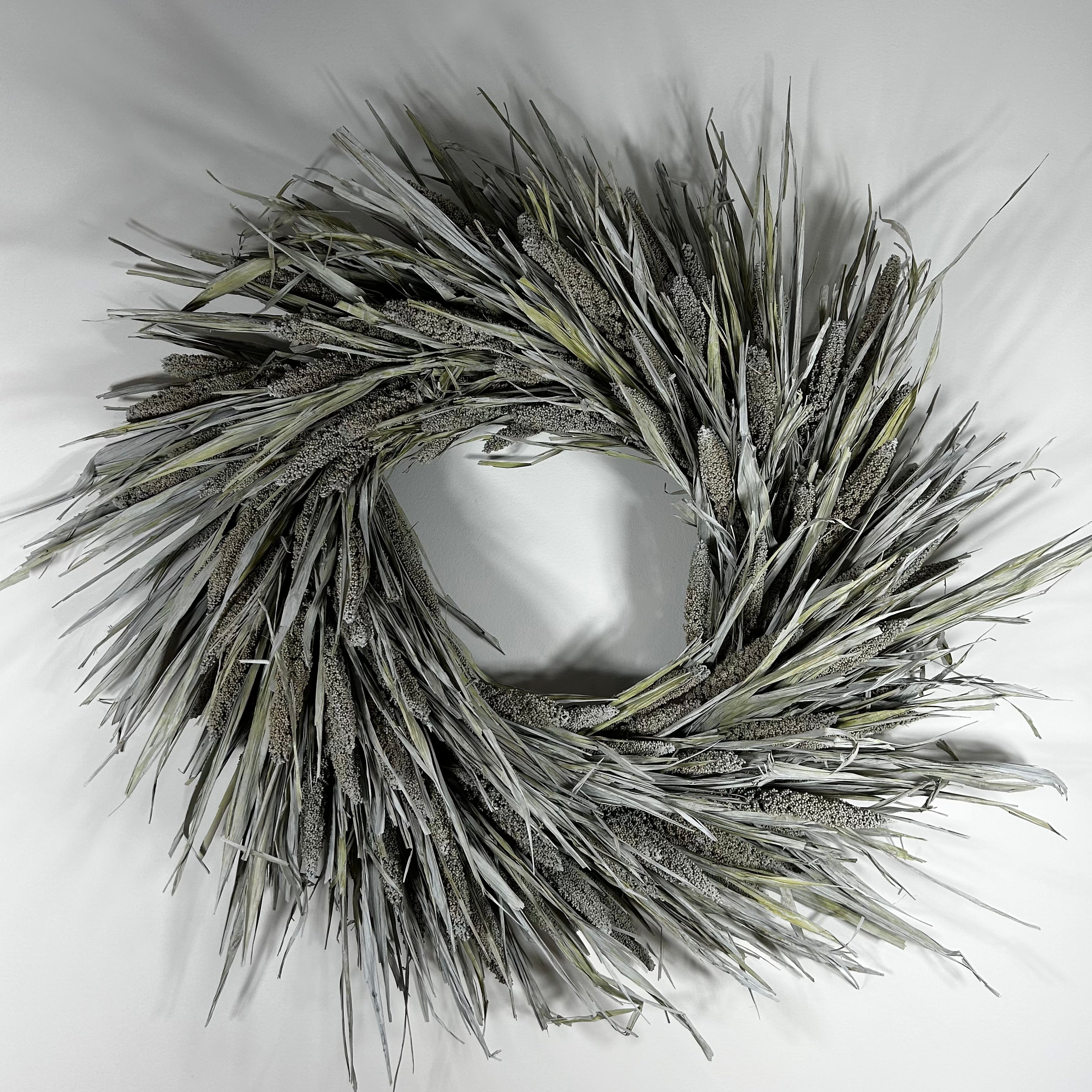 A circular decorative wreath made from dried reeds or cattails in muted gray, beige, and olive tones, photographed against a plain light background.