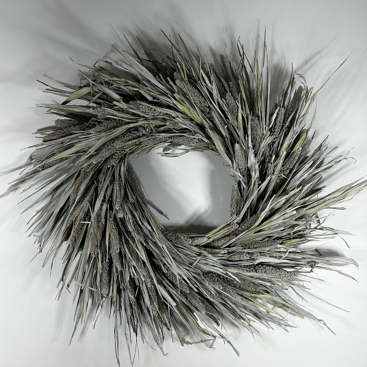 A circular decorative wreath made from dried reeds or cattails in muted gray, beige, and olive tones, photographed against a plain light background.