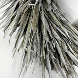 A close-up of a dried wheat or millet-style decorative wreath with silvery gray-green leaves and brownish seed clusters, photographed against a plain light background.