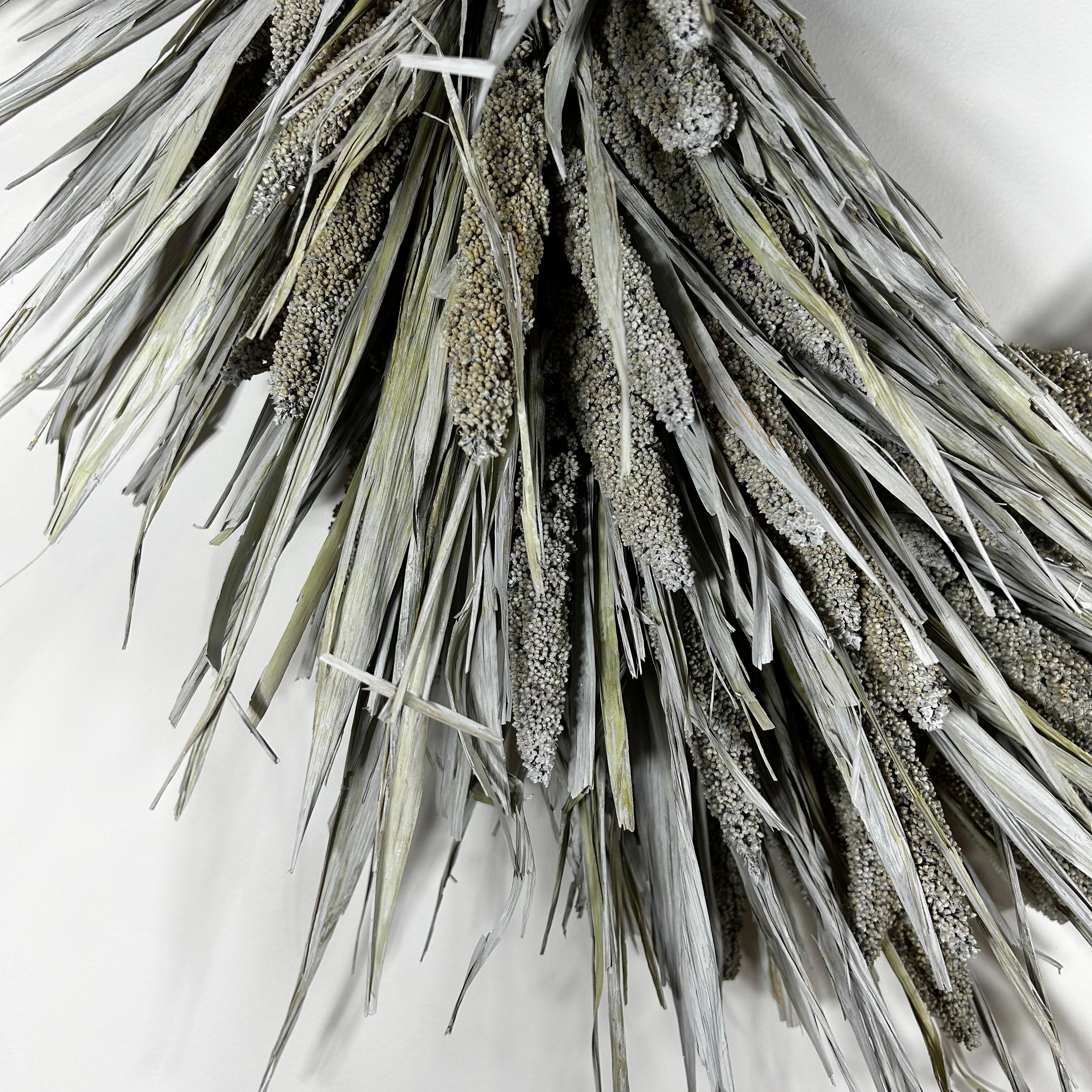 A close-up of a dried wheat or millet-style decorative wreath with silvery gray-green leaves and brownish seed clusters, photographed against a plain light background.