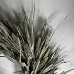 A dried floral arrangement featuring natural grain stalks and long, slender leaves in muted gray-green and beige tones, photographed against a plain light background.