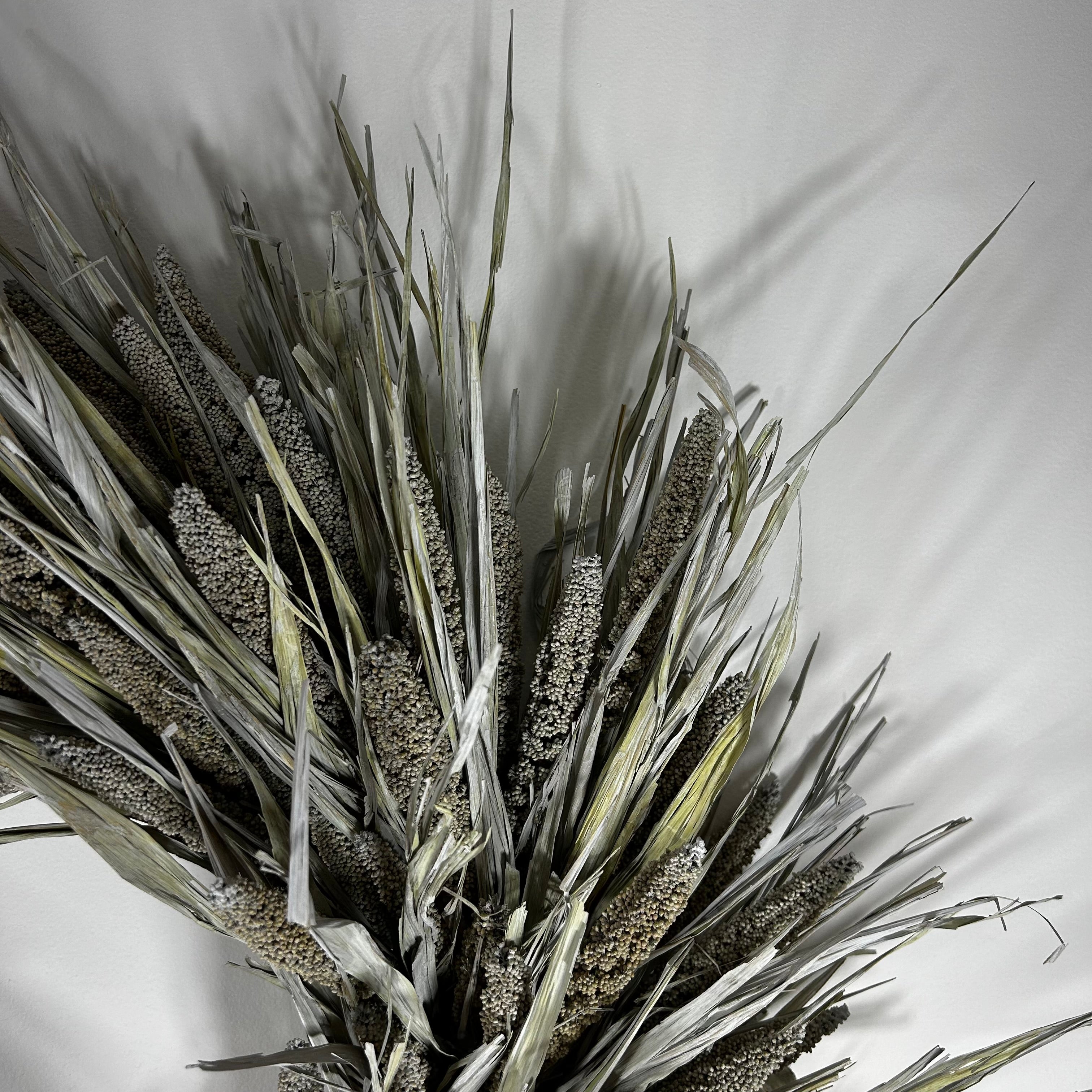 A dried floral arrangement featuring natural grain stalks and long, slender leaves in muted gray-green and beige tones, photographed against a plain light background.