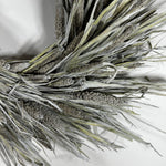 A close-up of a decorative faux wheat or millet wreath with textured silver-gray and olive-green dried grasses on a plain white background.