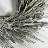 A close-up of a decorative faux wheat or millet wreath with textured silver-gray and olive-green dried grasses on a plain white background.
