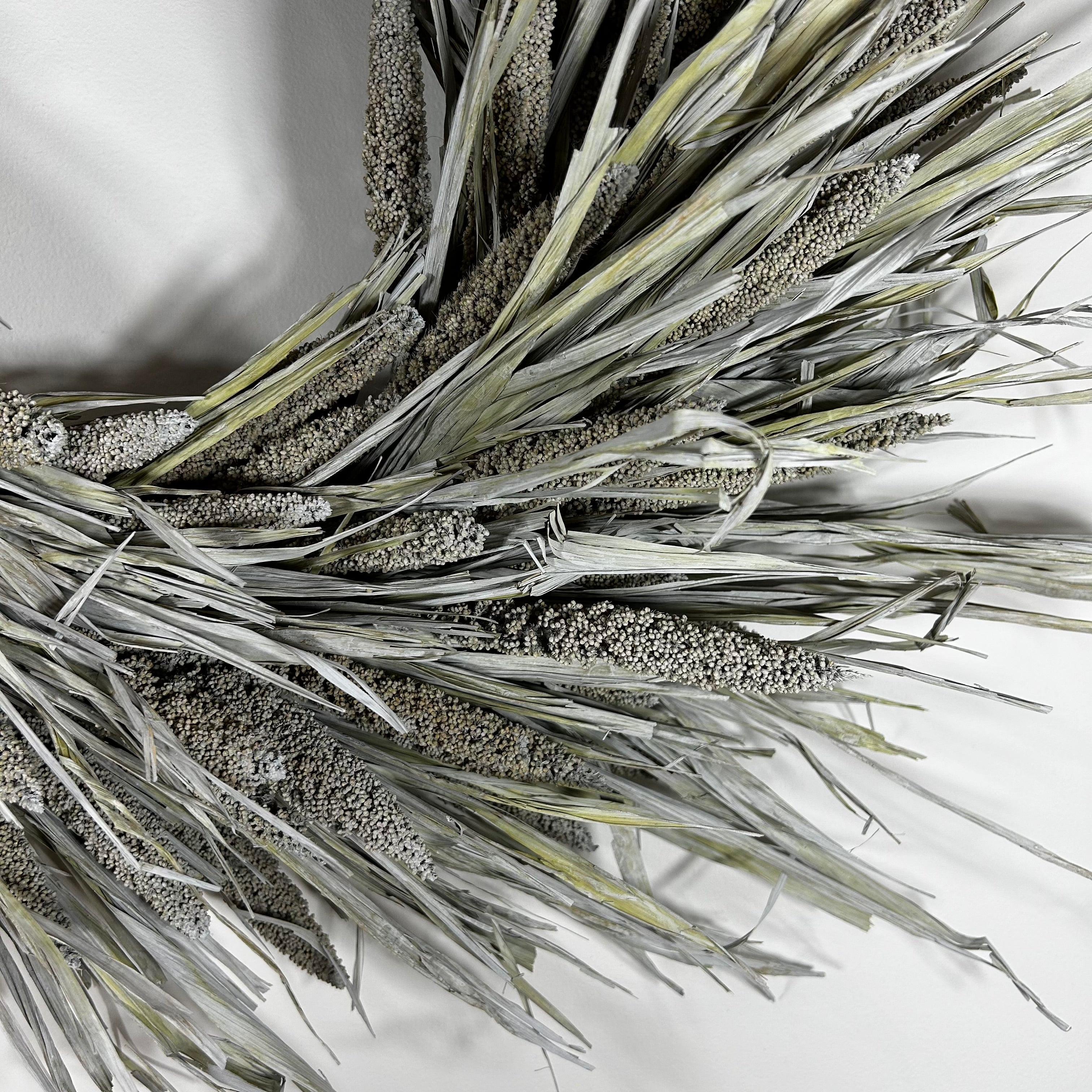A close-up of a decorative faux wheat or millet wreath with textured silver-gray and olive-green dried grasses on a plain white background.