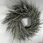 A natural reed wreath made of dried cattails and reeds, featuring textured brown seed heads and long gray-green leaves, displayed against a plain white background.