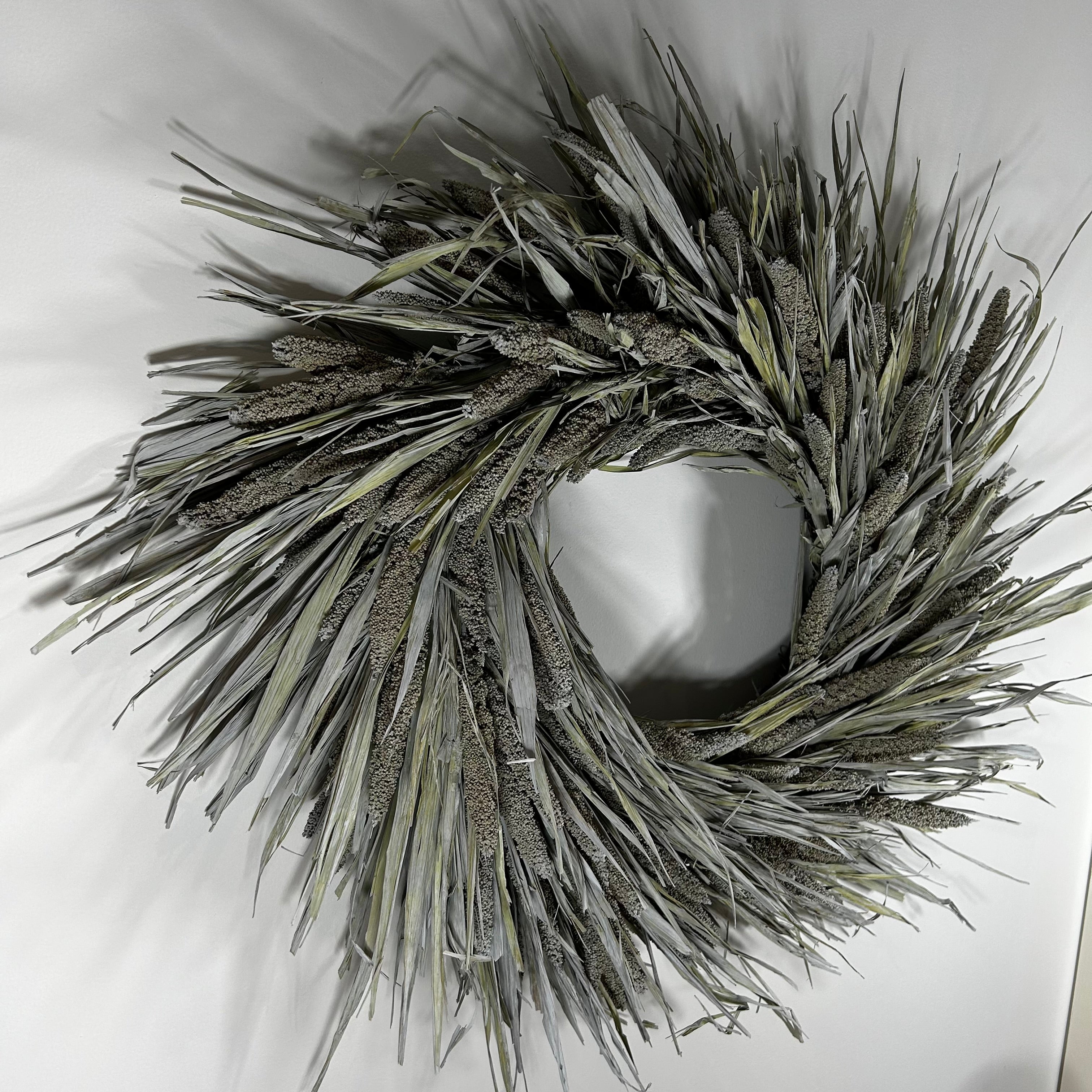 A natural reed wreath made of dried cattails and reeds, featuring textured brown seed heads and long gray-green leaves, displayed against a plain white background.