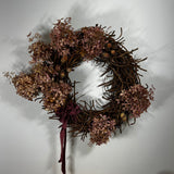 A rustic dried floral wreath with brown twig branches, dusty pink dried flowers, and a dark burgundy ribbon bow, hanging against a plain light gray wall.