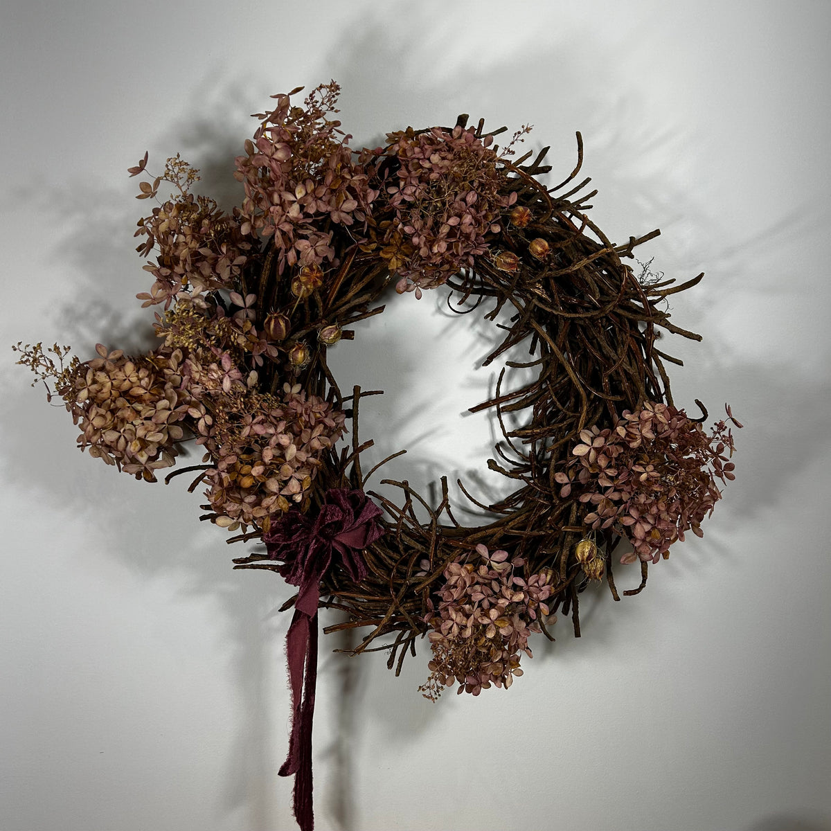 A rustic dried floral wreath with brown twig branches, dusty pink dried flowers, and a dark burgundy ribbon bow, hanging against a plain light gray wall.