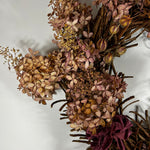 A dried floral arrangement featuring dusty pink and tan hydrangea-like blooms, brown twig branches, and a deep burgundy ribbon bow, photographed against a plain light background.