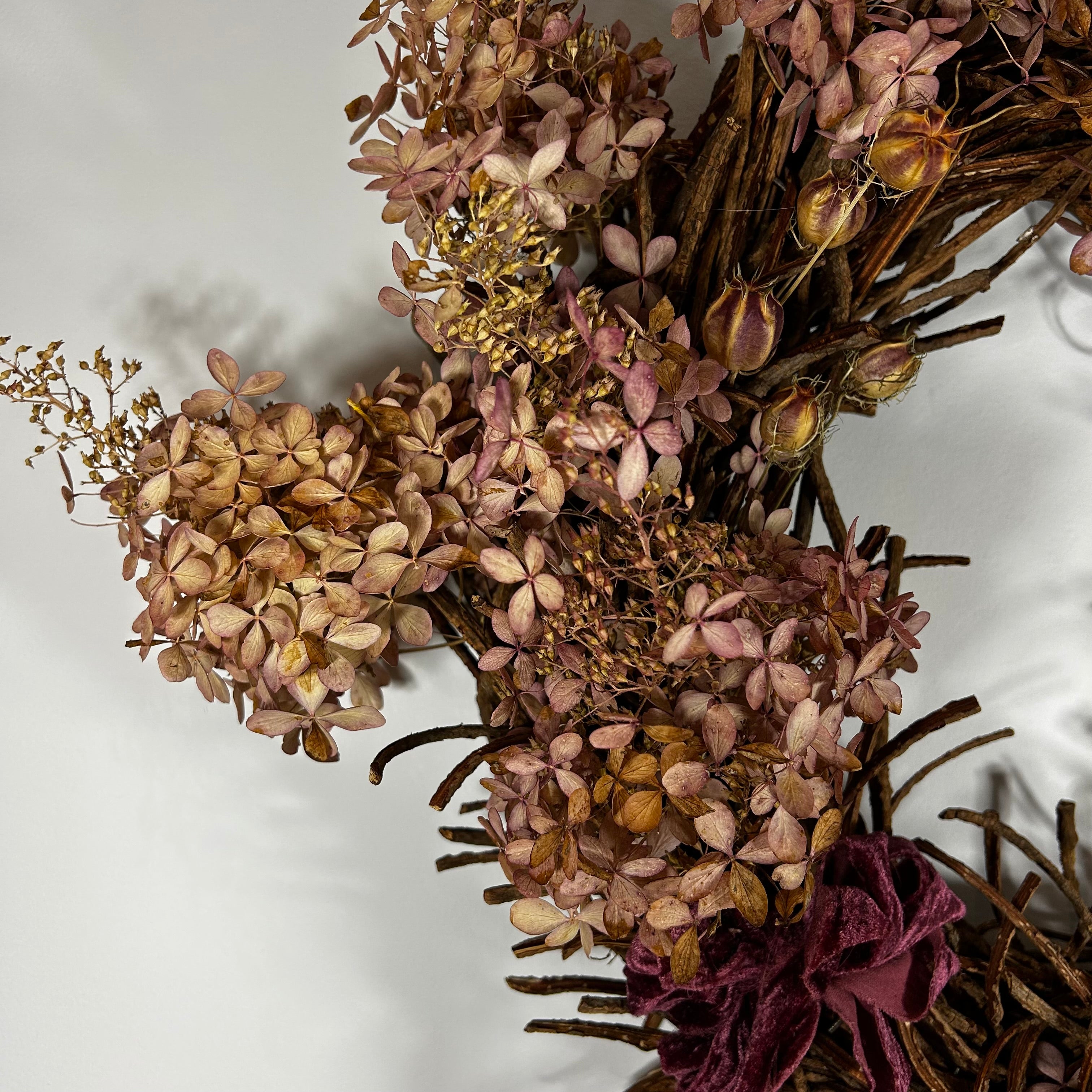 A dried floral arrangement featuring dusty pink and tan hydrangea-like blooms, brown twig branches, and a deep burgundy ribbon bow, photographed against a plain light background.