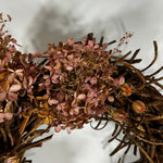 A close-up of a dried floral wreath featuring dusty pink hydrangea-like blossoms and a rustic brown twig base, photographed against a soft white background.