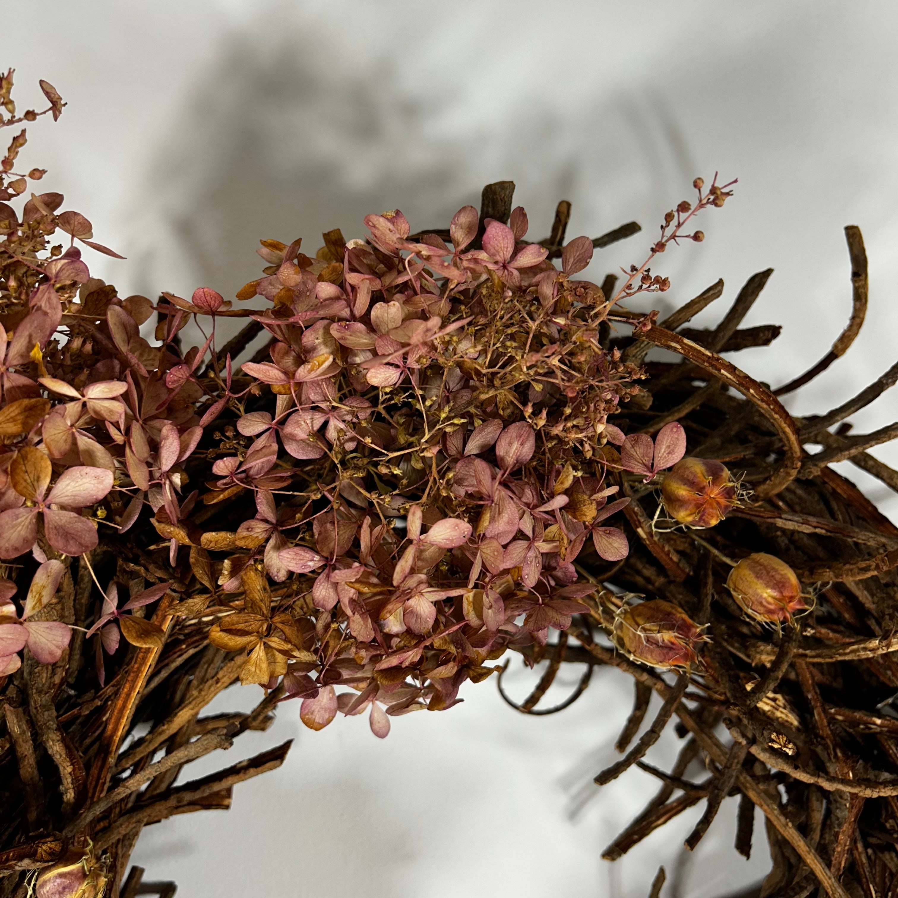 A close-up of a dried floral wreath featuring dusty pink hydrangea-like blossoms and a rustic brown twig base, photographed against a soft white background.
