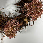 A rustic decorative wreath made of intertwined brown twigs and clusters of dried pinkish-brown flowers, photographed against a plain light background.