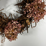 A rustic decorative wreath made of intertwined brown twigs and clusters of dried pinkish-brown flowers, photographed against a plain light background.