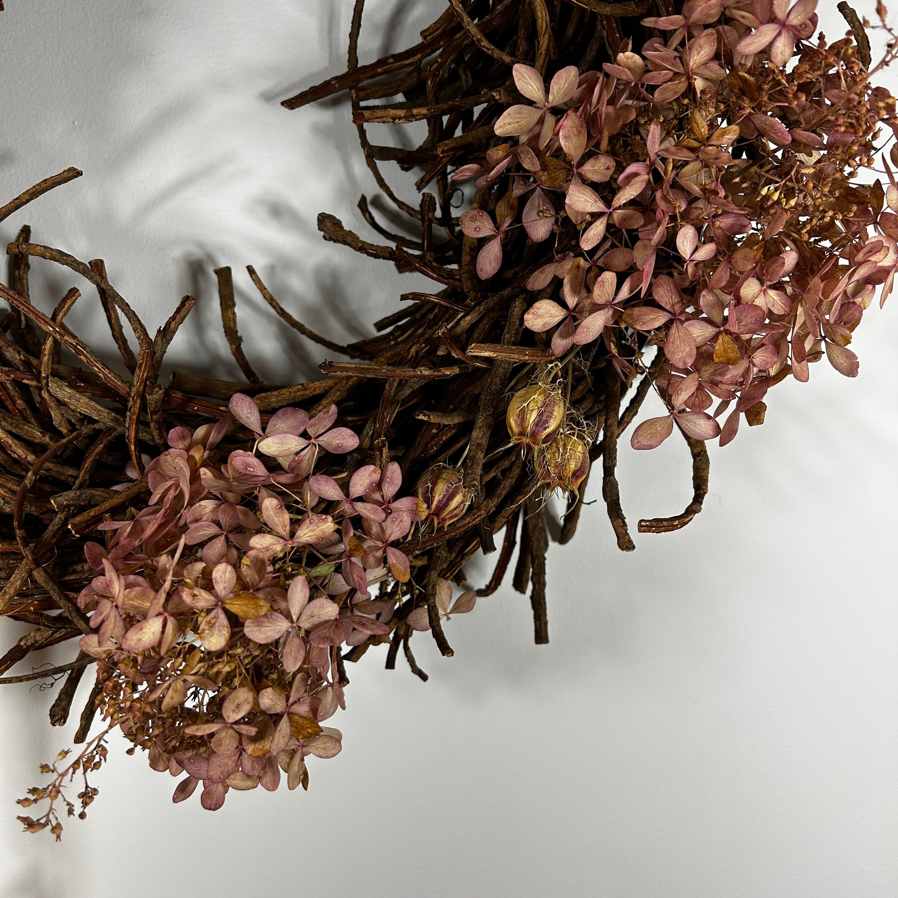 A rustic decorative wreath made of intertwined brown twigs and clusters of dried pinkish-brown flowers, photographed against a plain light background.