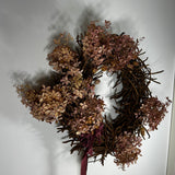 A rustic dried floral wreath made of brown twig branches and dusty mauve-pink hydrangea blooms, with a dark burgundy ribbon at the bottom, hanging against a plain white wall.