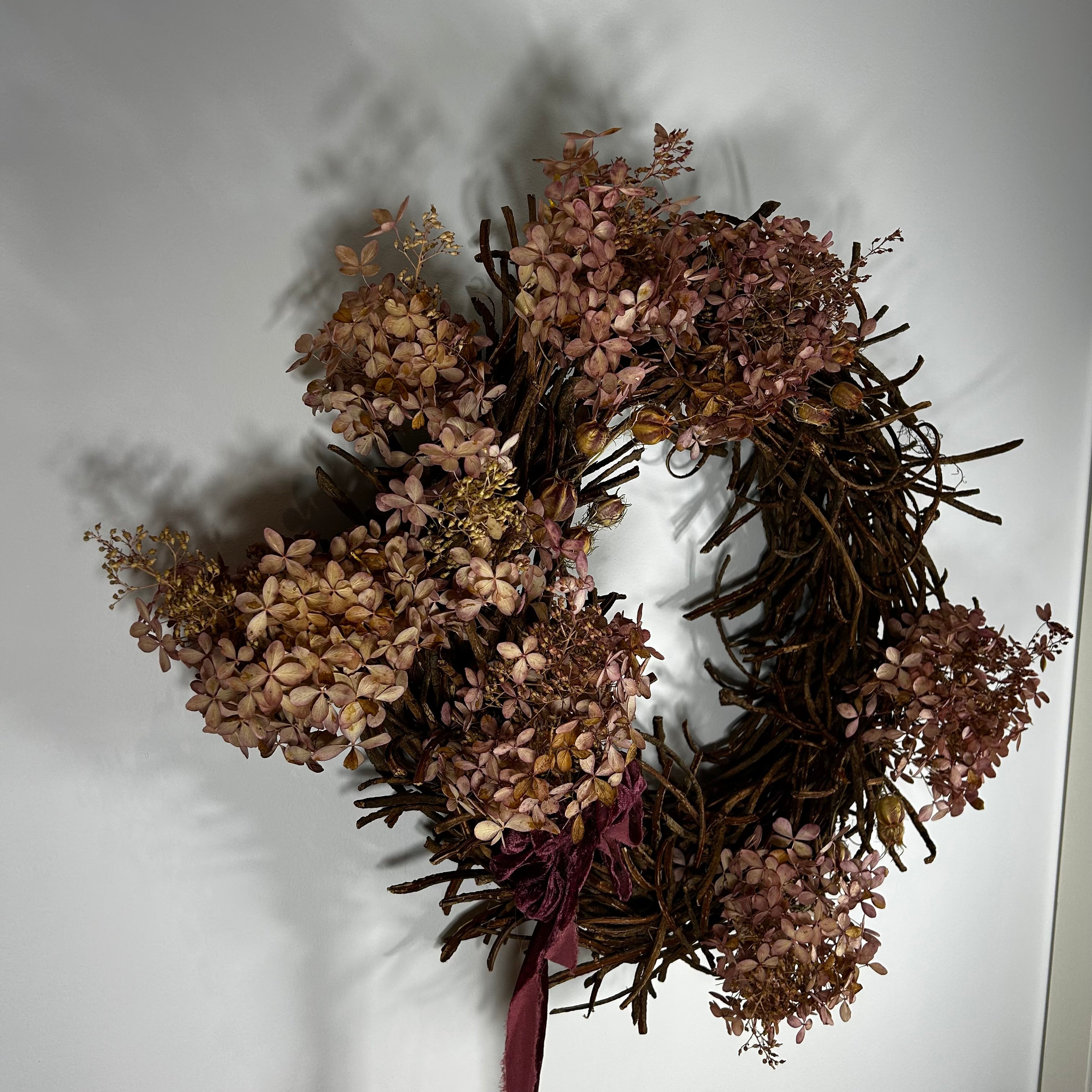 A rustic dried floral wreath made of brown twig branches and dusty mauve-pink hydrangea blooms, with a dark burgundy ribbon at the bottom, hanging against a plain white wall.