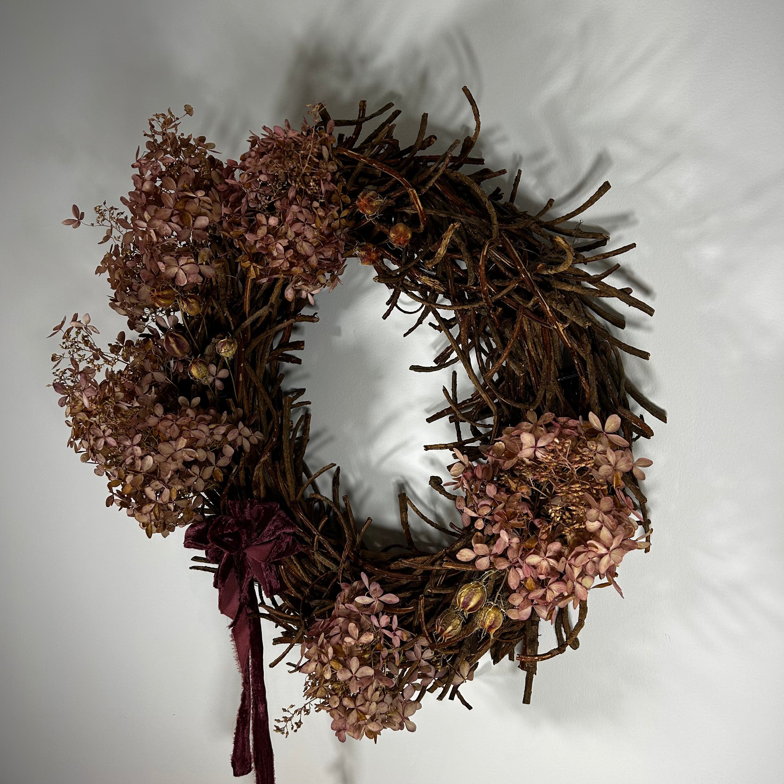 A rustic dried flower wreath made of brown twig branches, decorated with dusty pink hydrangea blooms and a dark burgundy ribbon bow at the bottom left, shown against a plain off-white background.