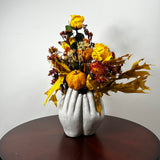 An autumn floral arrangement in a sculptural gray vase shaped like hands, displayed on a dark round wooden table against a plain light gray wall. The arrangement features yellow roses, a small orange pumpkin, red berries, and golden maple leaves.