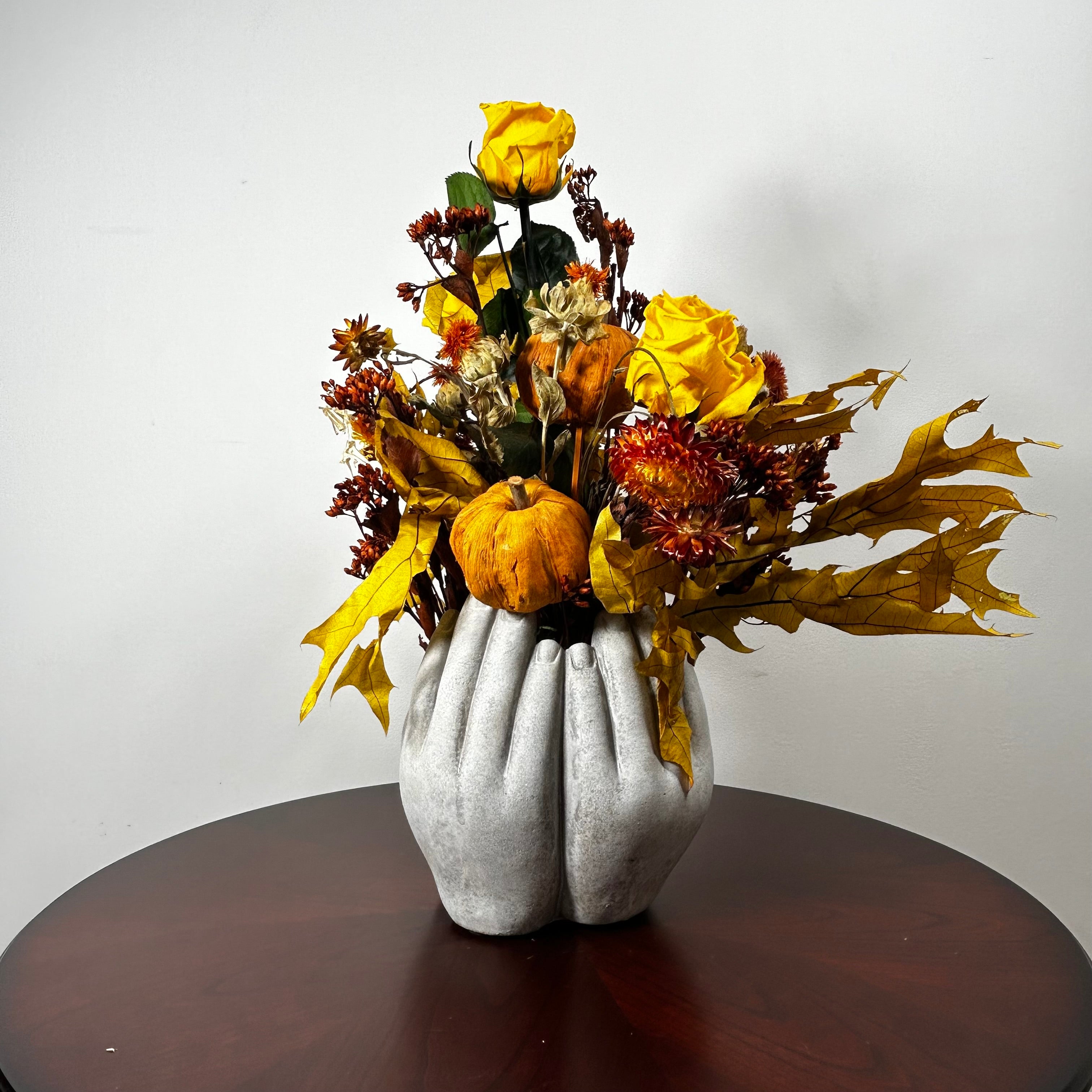 An autumn floral arrangement in a sculptural gray vase shaped like hands, displayed on a dark round wooden table against a plain light gray wall. The arrangement features yellow roses, a small orange pumpkin, red berries, and golden maple leaves.
