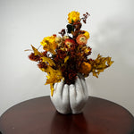 A decorative arrangement of dried yellow, orange, and brown flowers with autumn leaves in a sculptural white vase shaped like two hands, displayed on a round dark wooden table against a plain light gray wall.