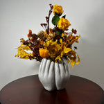 An autumn floral arrangement featuring yellow roses, orange pumpkins, and dried leaves in a sculptural white vase shaped like two hands, displayed on a dark wooden table against a plain light gray wall.