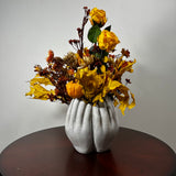 An autumn floral arrangement featuring yellow roses, orange pumpkins, and dried leaves in a sculptural white vase shaped like two hands, displayed on a dark wooden table against a plain light gray wall.