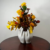 An autumn floral arrangement with yellow and orange faux leaves and flowers in a sculptural pair of white hands vase, displayed on a dark wooden table against a plain light gray wall.