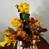 An autumn-themed artificial floral arrangement featuring yellow roses, orange and red berries, yellow leaves, and green foliage, displayed in a white fabric vase on a dark wooden surface against a plain light gray wall.